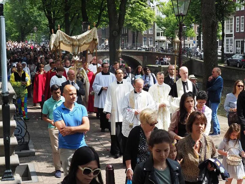 Multitud de fieles participa en una procesión de Corpus Christi por las calles de una ciudad europea, acompañando al Santísimo Sacramento bajo palio.