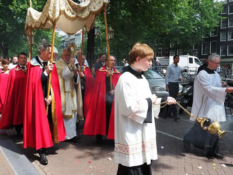 Procesión del Corpus Christi con sacerdotes y fieles llevando el Santísimo Sacramento bajo palio por una calle urbana.