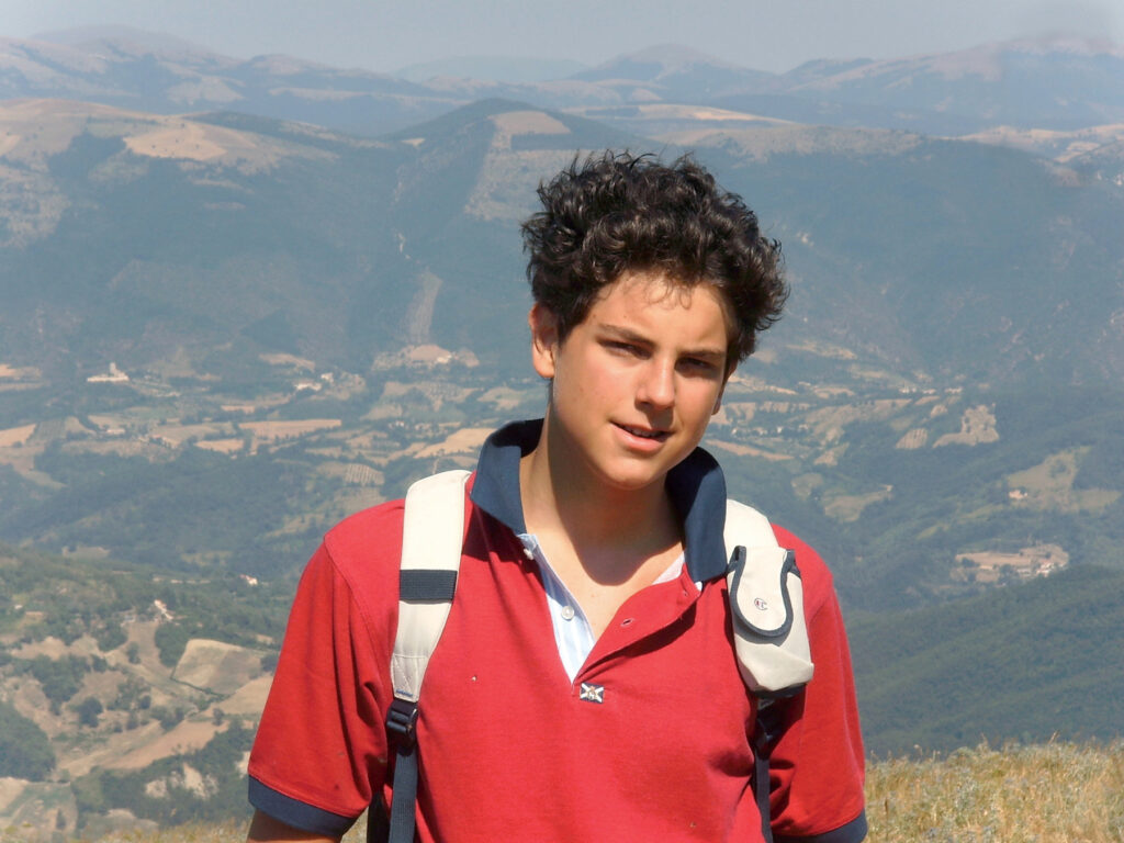 El joven san Carlo Acutis en una foto al aire libre con un paisaje de montañas al fondo, vistiendo un polo rojo y una mochila.