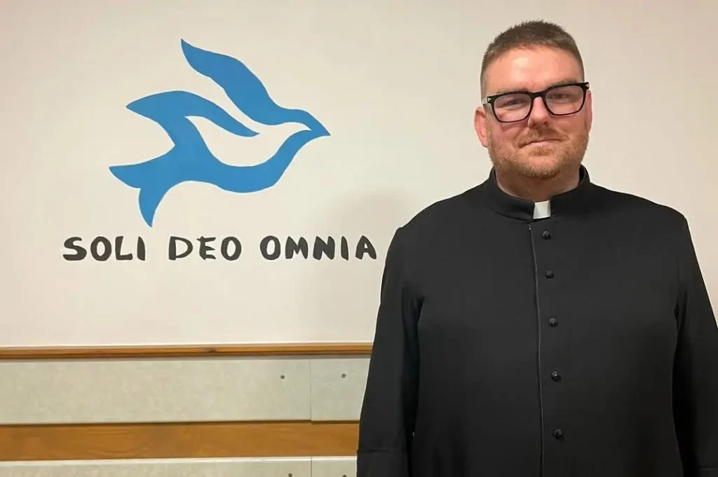 Krisztof Patejuk, prillidega, musta kasuka ja Rooma kraega riietatud, seisab valge seina ees, millel on sinine tuvilogo ja tekst "SOLI DEO OMNIA".