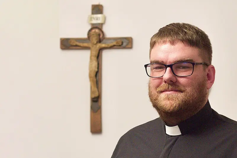 Sacerdote cattolico polacco con colletto e occhiali, in piedi accanto a un crocifisso di legno appeso alla parete.