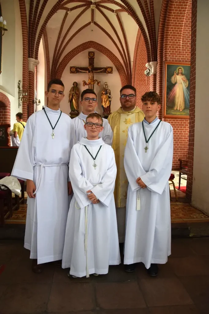Neli last ja poola preester, kes on riietatud valgetesse ja kuldsetesse liturgilistesse rõivastesse kirikus. Lastel on ristid kaelas ja preestril stoola. Taustal on telliskivikaared ja altar koos ristiga.