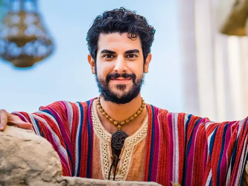 El actor Joey Vahedi interpretando al apóstol Tomas en la serie The Chosen, vestido con una túnica a rayas rojas y beige, mira directamente a la cámara y sonríe.
